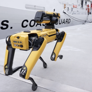 A four-legged (quadrupedal) robot walks on a dock next to a U.S. Coast Guard ship.