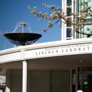 The exterior main entrance of MIT Lincoln Laboratory is shown with a radar antenna in the background.