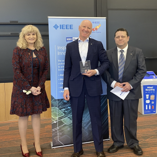Two IEEE staff stand to the left and right of Laboratory staff member Jeffrey Herd as he receives his award, a glass plaque. 