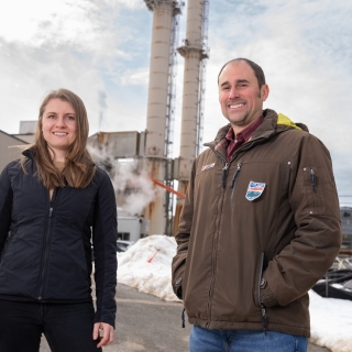 Jean Sack and Chris Lashway pose for a photo outside, with power-plant infrastructure in the background.