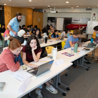 LLCipher students sit in groups of two at long tables working on laptops. Two instructors are helping the students.