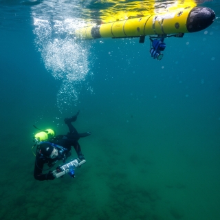 A diver carrying a tube-shaped tablet and an autonomous underwater vehicle swim underwater together.