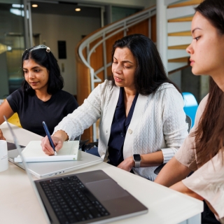 Three people sit at a desk with laptops and a notepad.