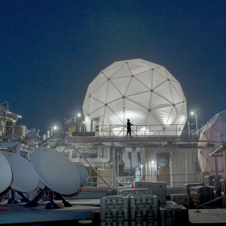 Four satellite dishes and two radomes, each covering satellite communications antennas, are shown on the roof of Lincoln Laboratory among other equipment.