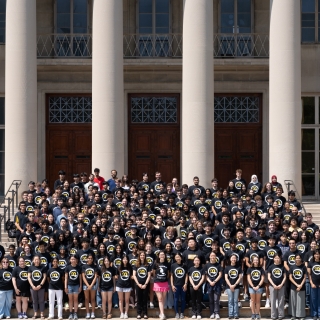 BWSI students wear matching black shirts with the BWSI logo and stand on steps outside of an MIT building with columns in the background. 
