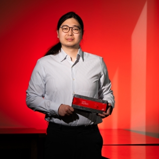 A photo of Jeffery Lim against a red background, holding a hardware platform (rectangle, about 4 by 12 inches). 