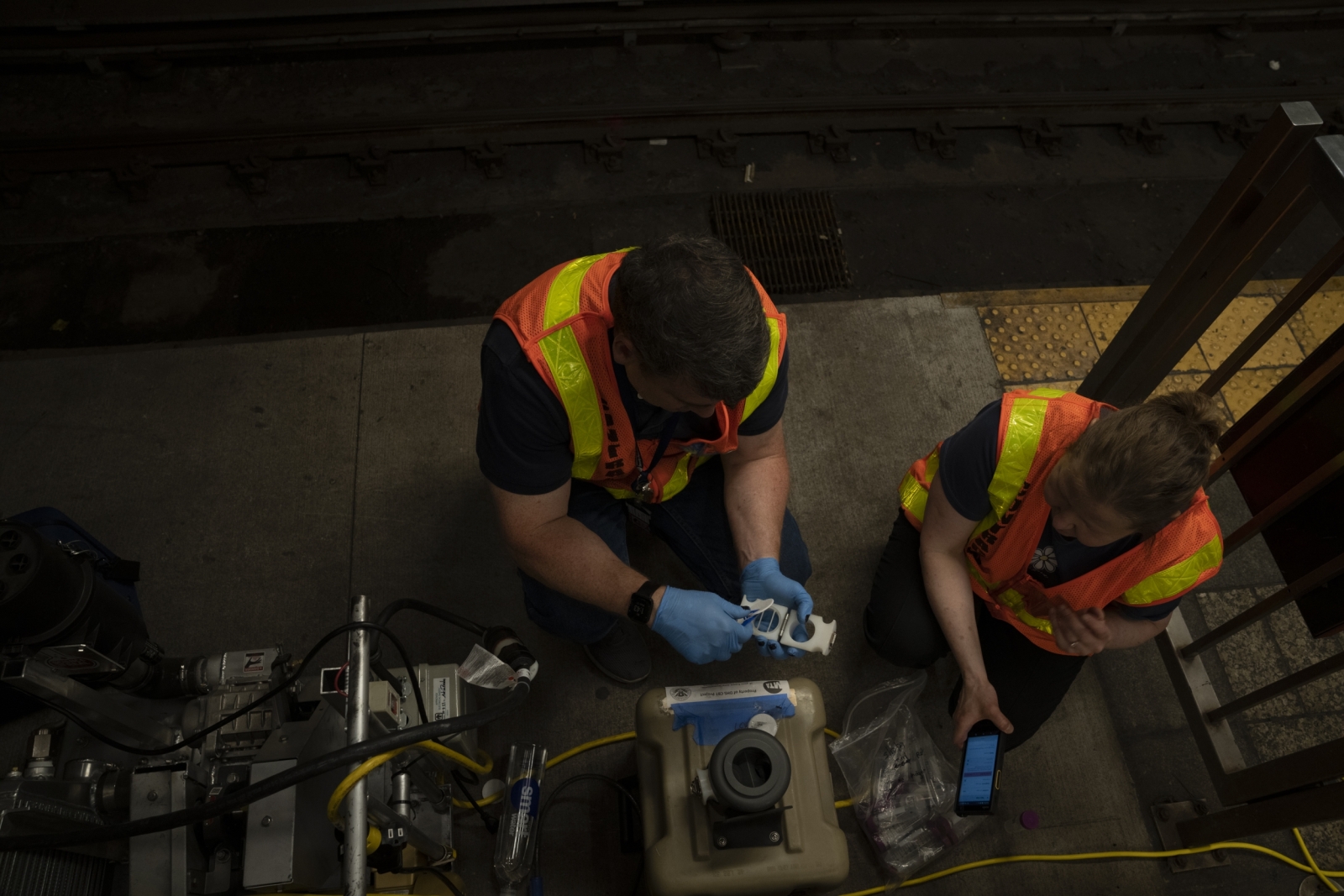 Two Laboratory staff members in orange vests kneel in front of a sensor on a subway platform to collect data from a recent test. 