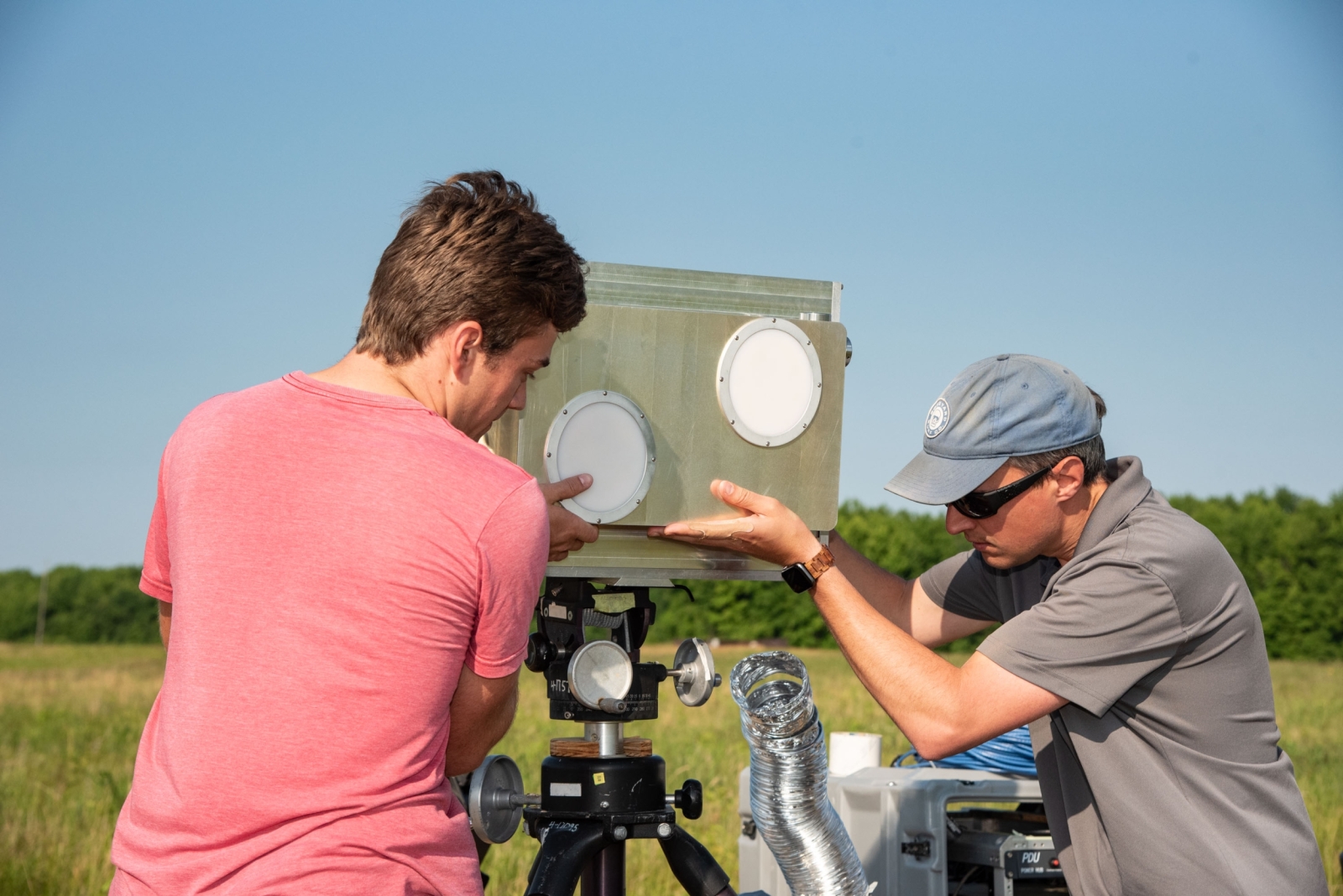 Two men adjust a metal antenna unit, approximately 12 in by 18 in, on a tripod 