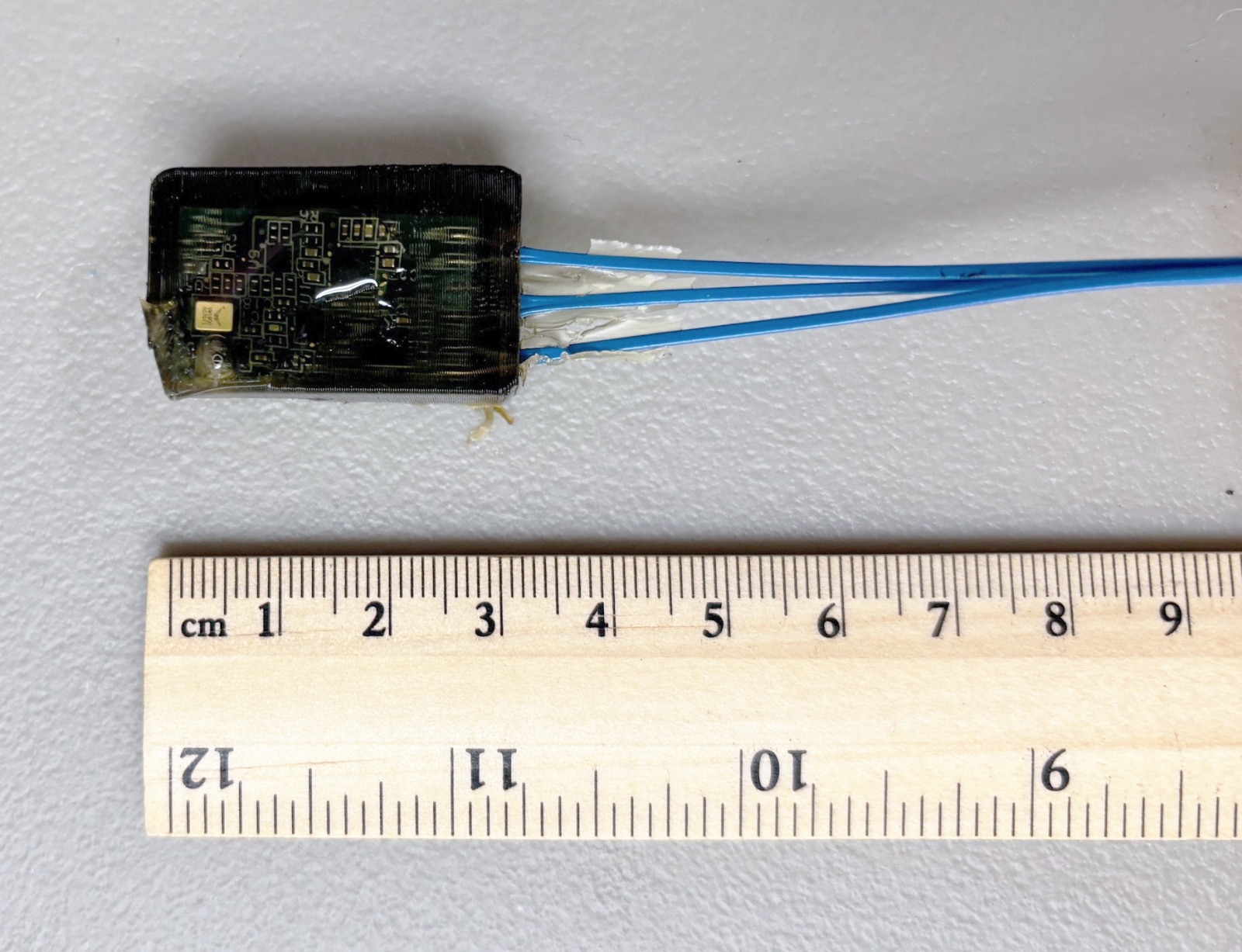 A photo of a small electronic device with blue wires, next to a ruler for scale. The device is about 4 centimeters long. 