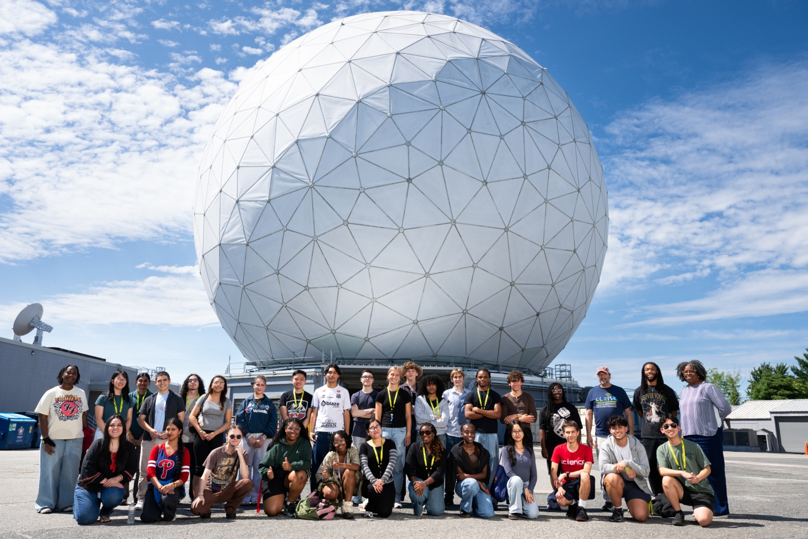 The full 2026 LLRISE class takes a group photo in front of the white dome of the Haystack radar with a blue sky and clouds in the background. 