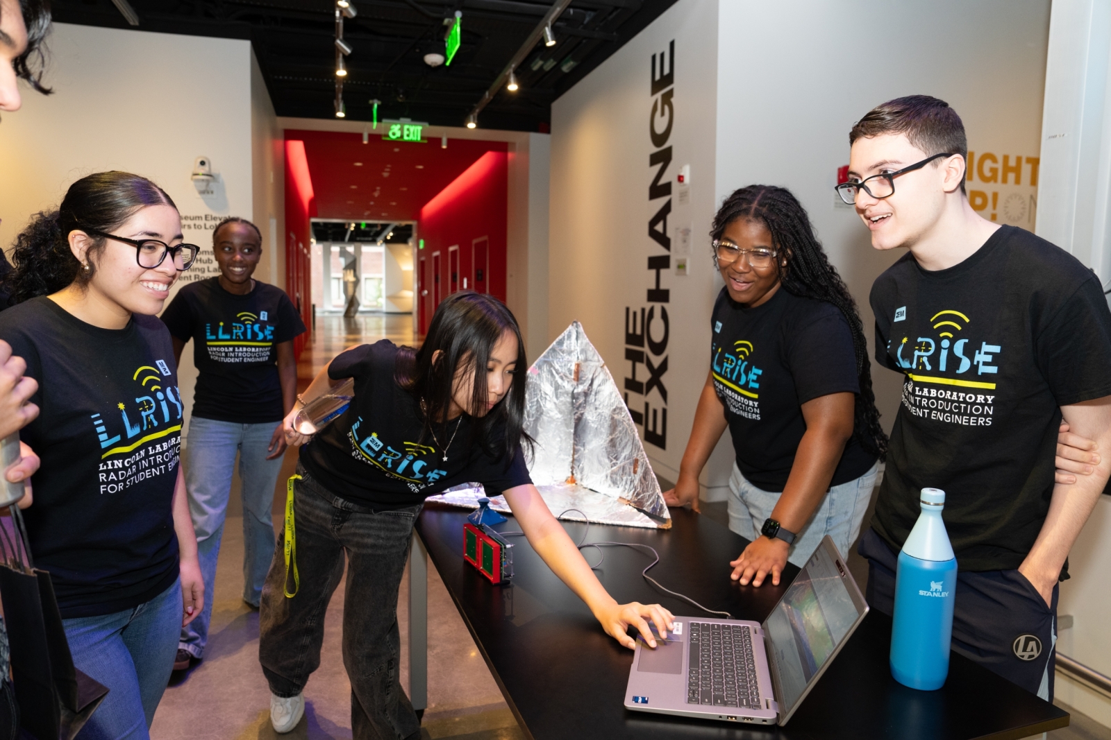 A group of LLRISE students wearing LLRISE shirts smile while preparing equipment inside of the MIT Museum.