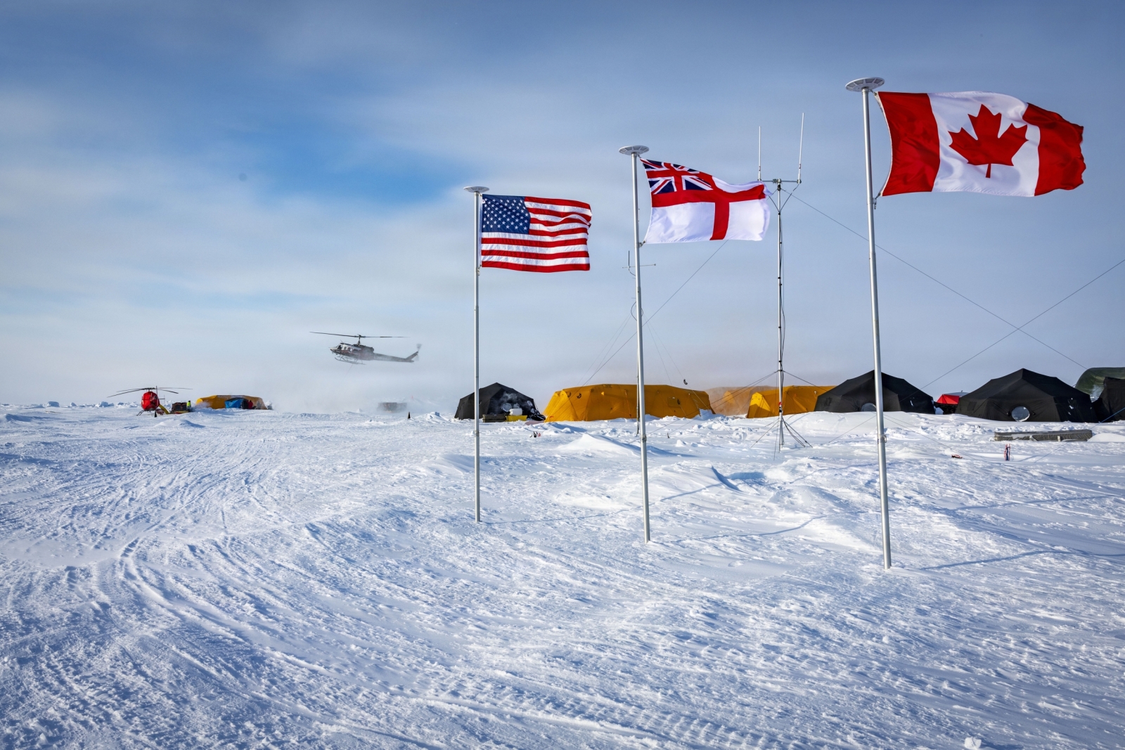 Multiple tents sit on the Arctic sea ice, with flags of the United States, United Kingdom, and Canada planted in the ice. 