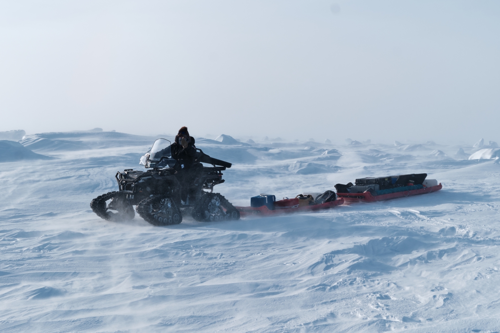 A person drives 4x4 vehicle with tracks pulling a sled with equipment.