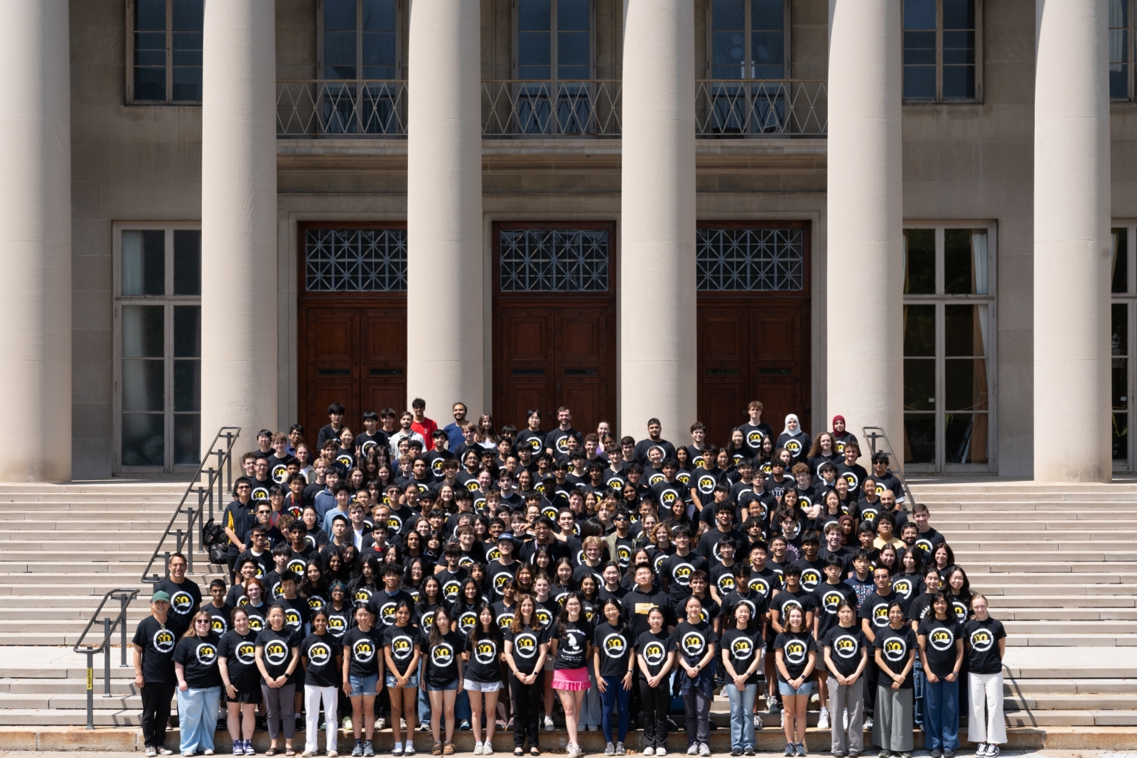 BWSI students wear matching black shirts with the BWSI logo and stand on steps outside of an MIT building with columns in the background. 