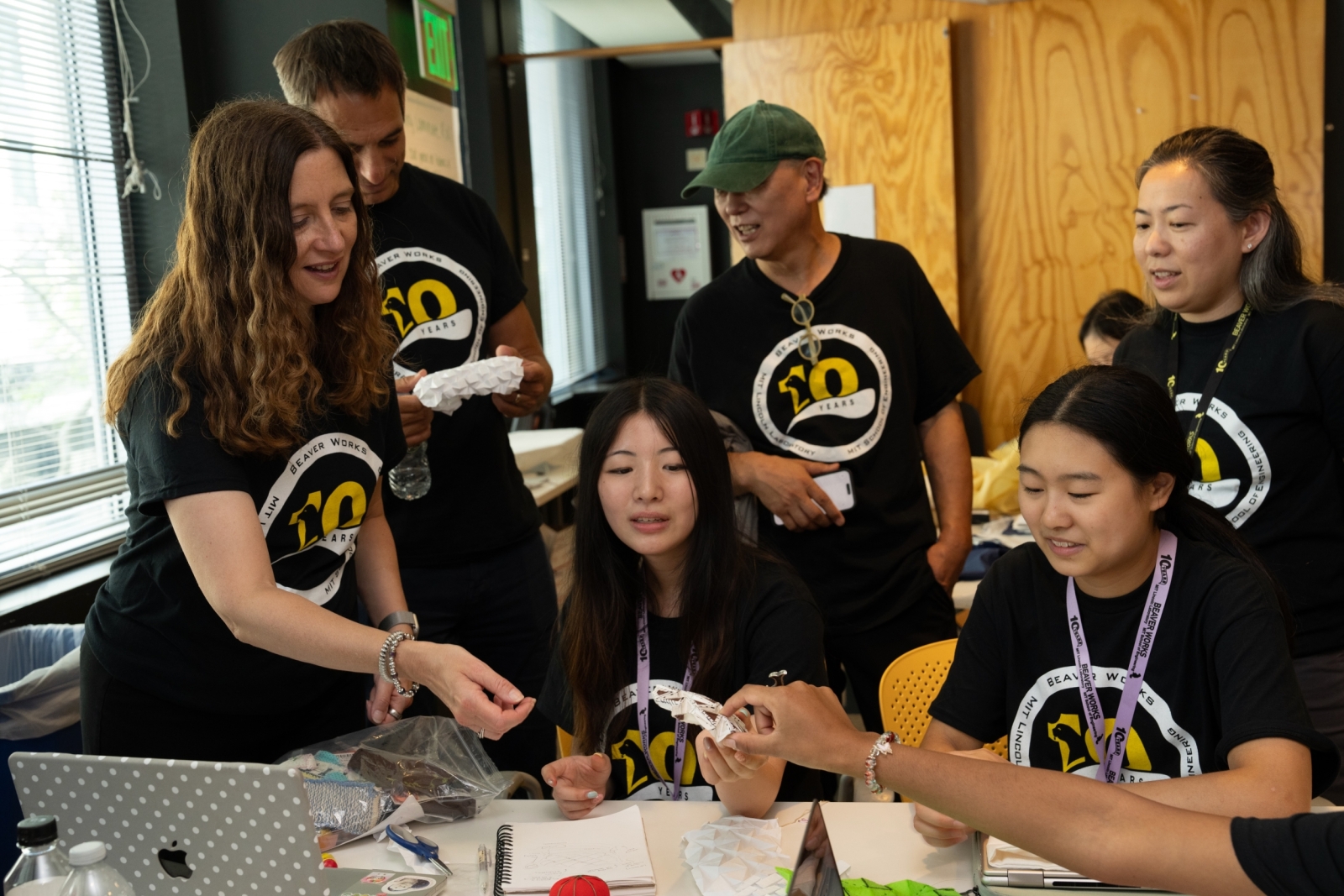 Laboratory Director Melissa Choi interacts with students at a table filled with project items. All wear a BWSI black shirt with logo. 