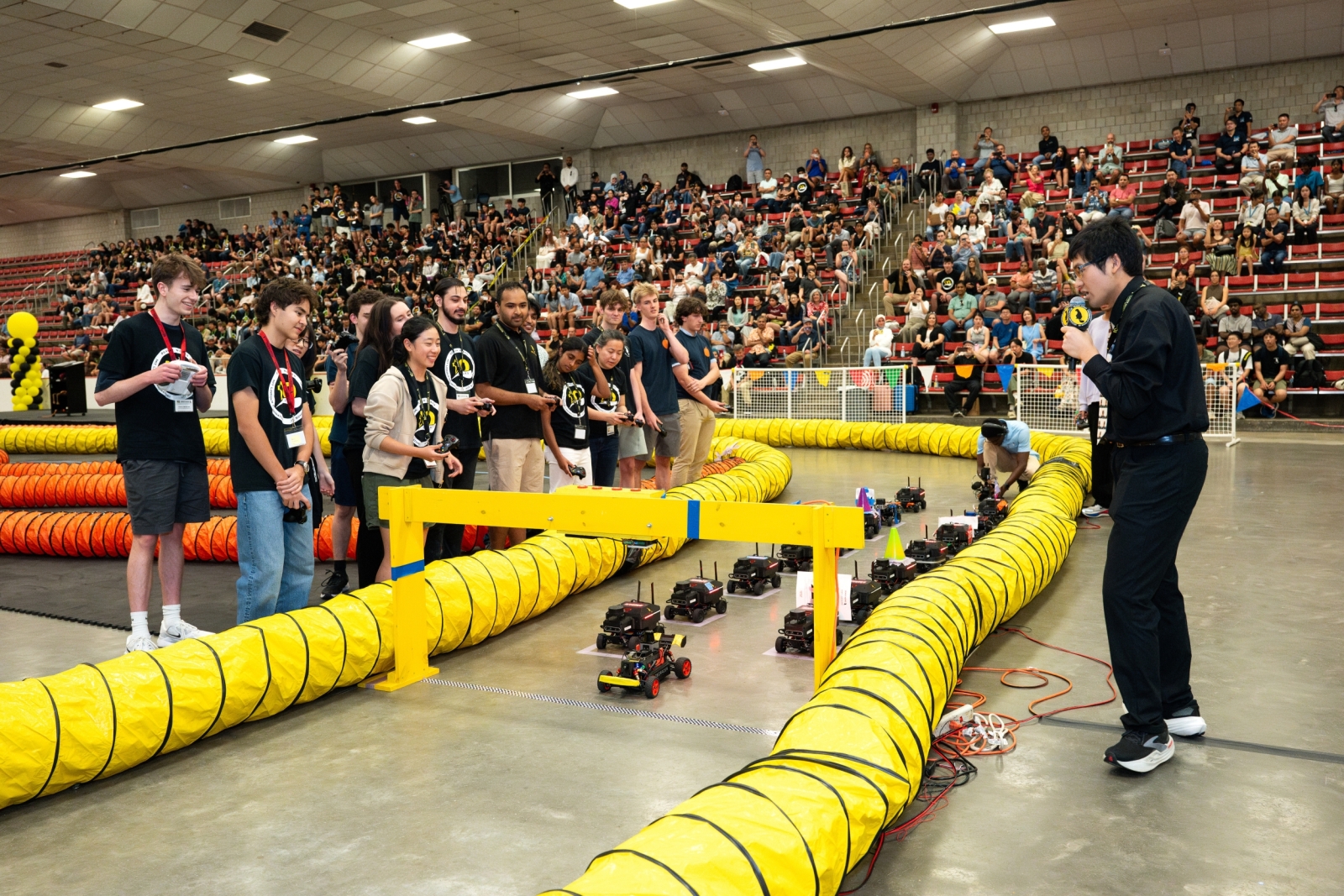 Race participants stand behind yellow tubing that denotes a racetrack inside an indoor arena. The racecar vehicle entries are gathered at a starting point while spectators watch on bleachers in the background. 