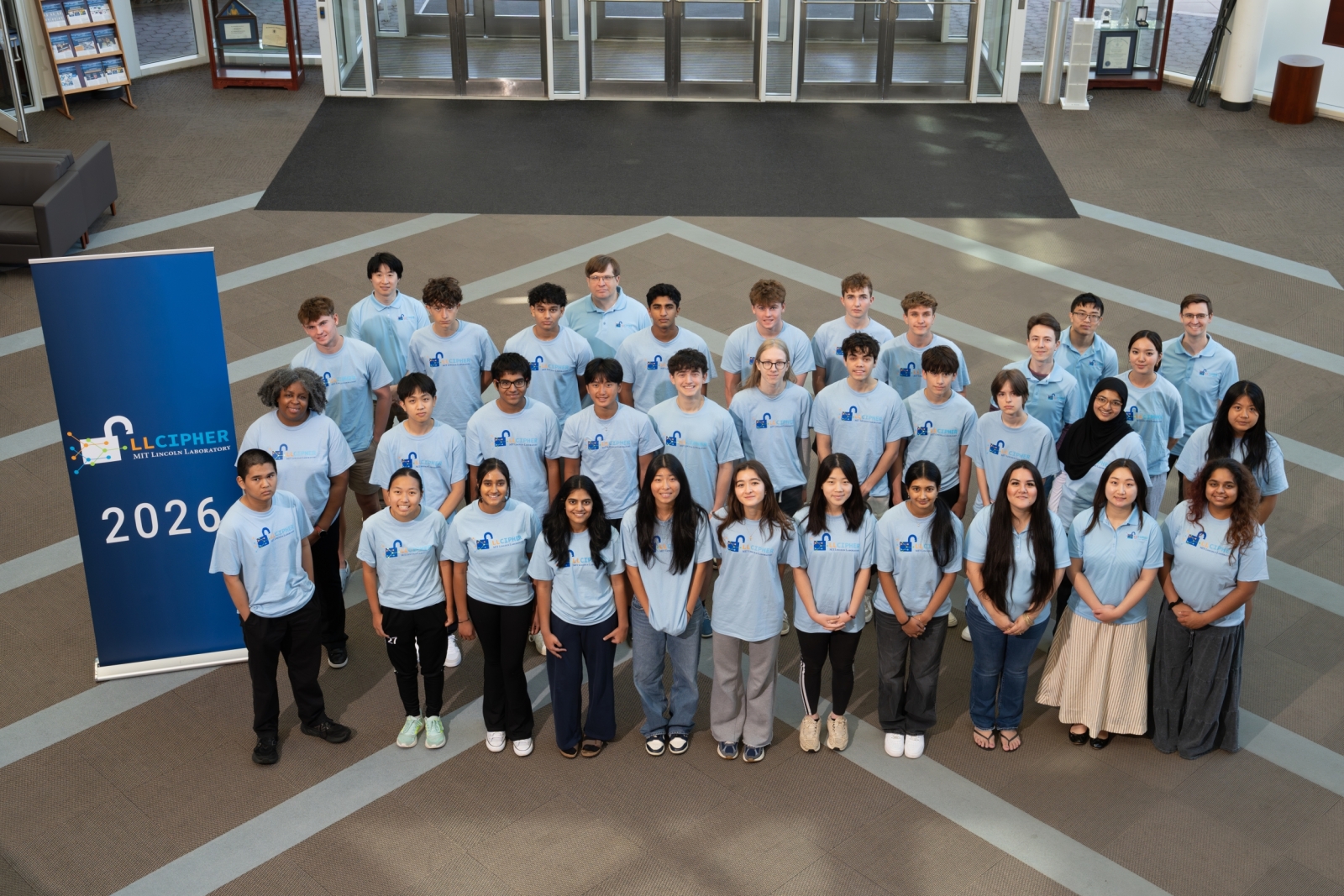 The year's LLCipher students smile for a photo in the Laboratory's main entrance, all wearing light blue LLCipher T-shirts. 
