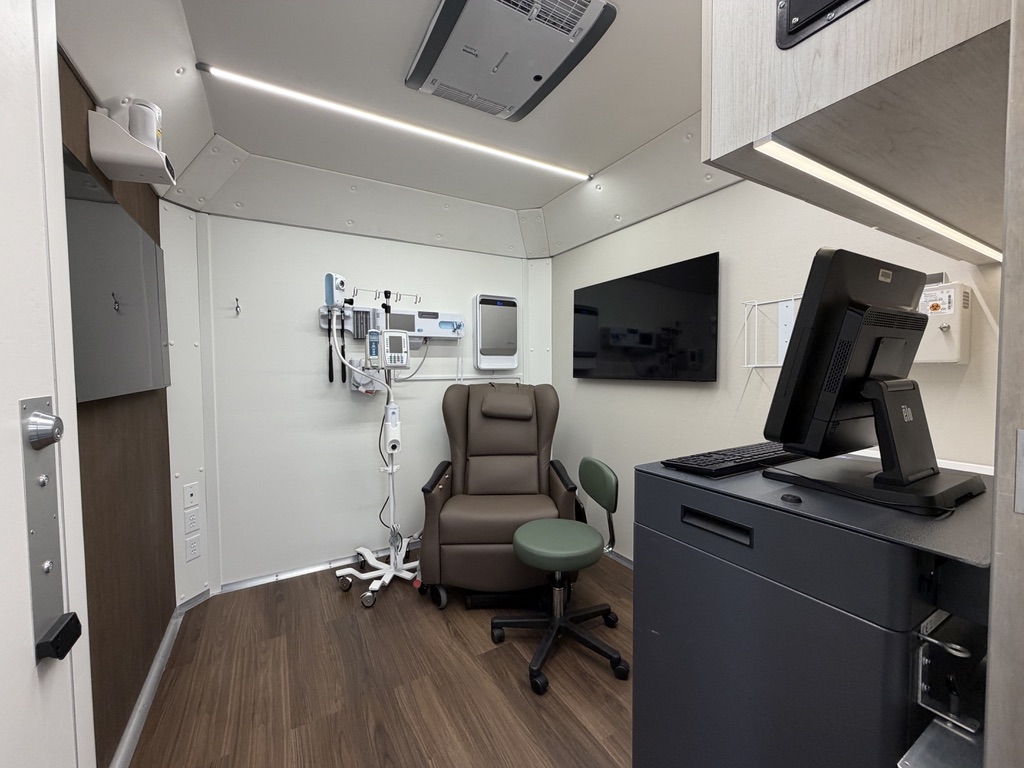 A photo of the interior of the Mission Mobile Medical vehicle showing the patient treatment area that has a chair in the center and a monitor, computer stand, and desk to the right. 