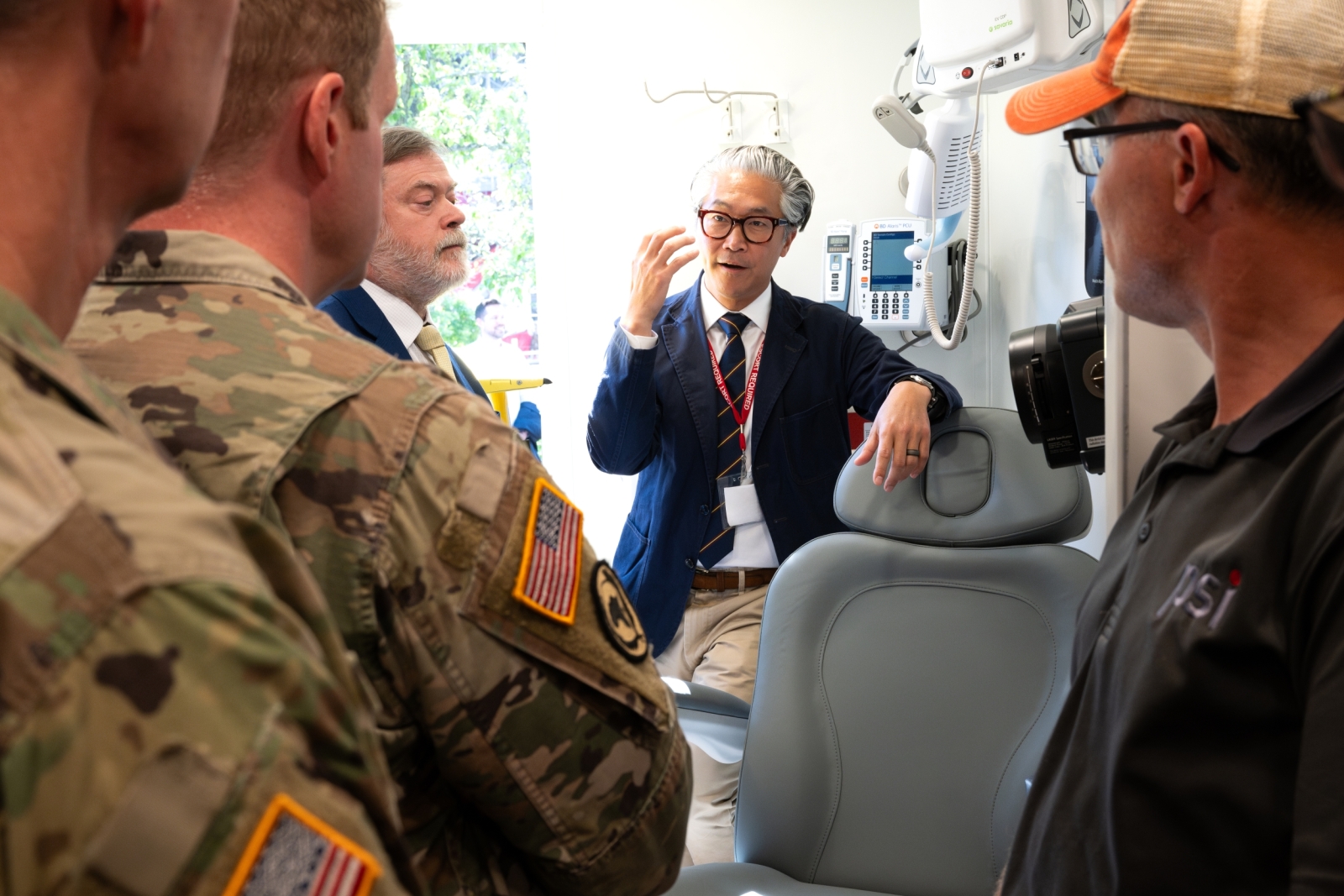 ARPA-H program manager Bon Ku faces the camera while talking to service members who are in the foreground. Part of the PSI vehicle's patient chair setup is visible next to Ku. 