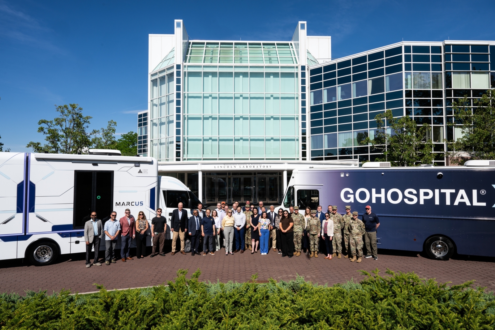 A group photo of the research teams, Laboratory personnel, and visiting soldiers at the Laboratory's front entrance with the two vehicles behind them. 
