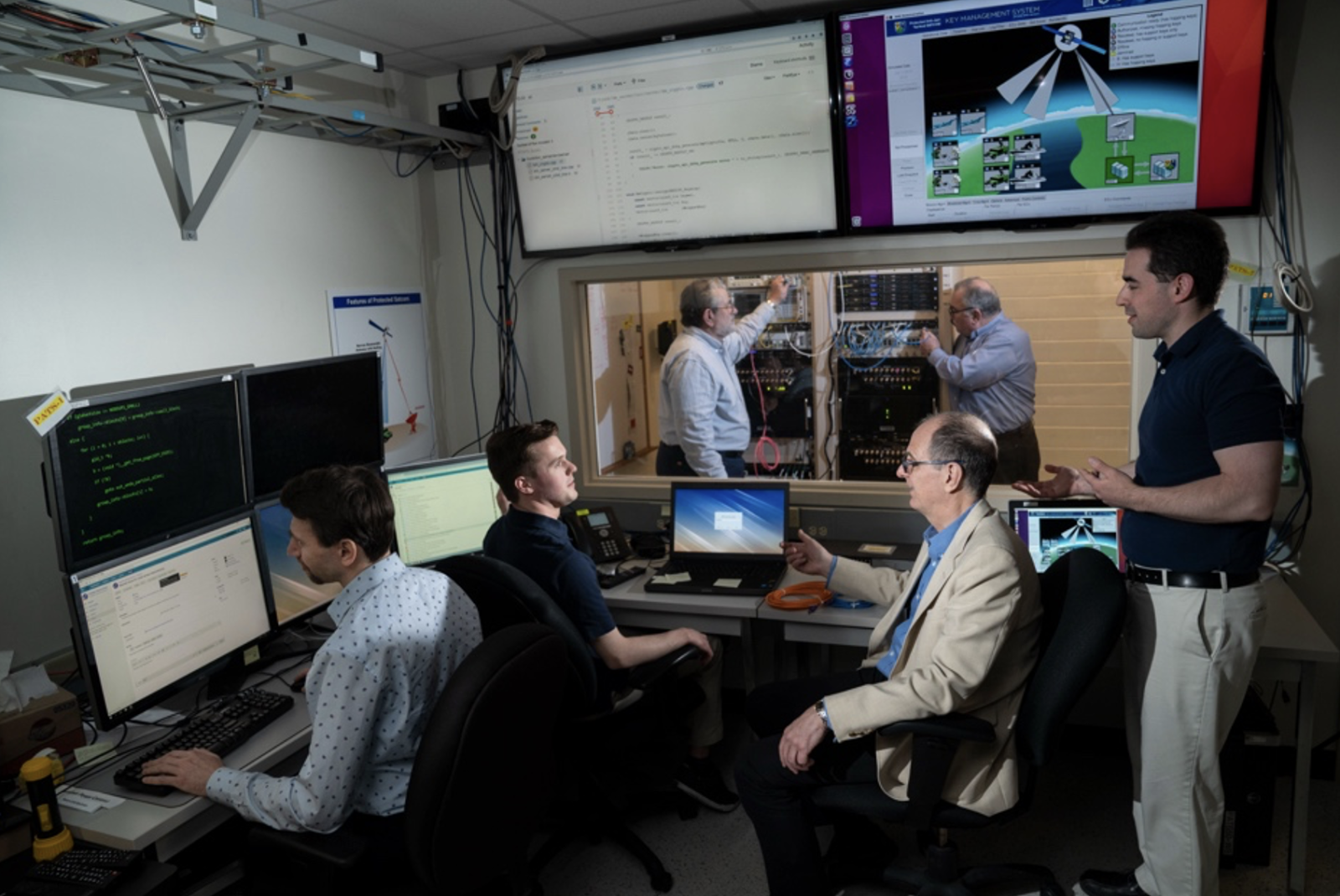 Six researchers stand in a lab, showing computer screens, monitors, and a rack of hardware. 