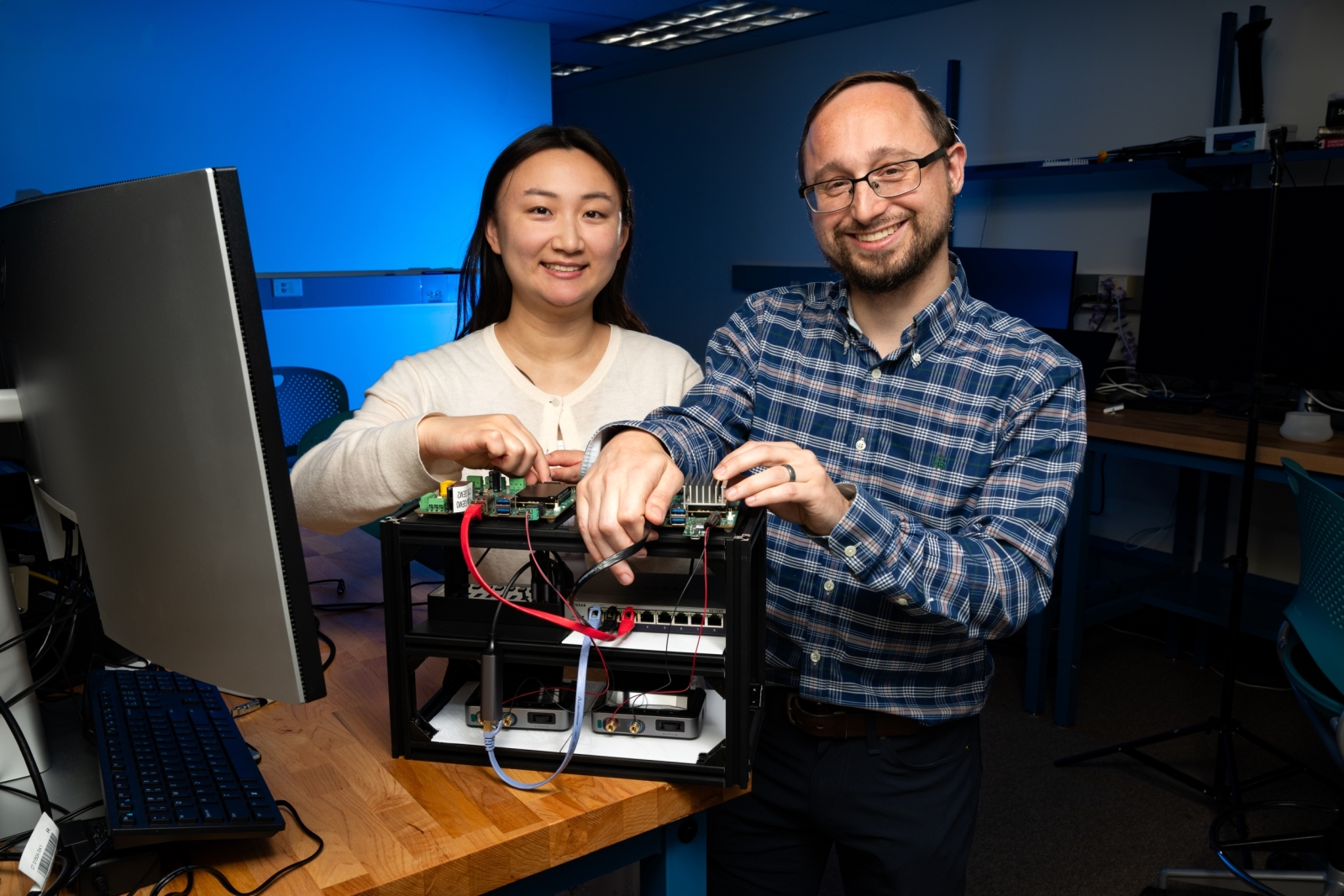 Andrea Lin and Samuel Jero smile for a photo inside of a lab space. They are handling an approximately 2 ft X 1 ft X 1ft hardware rack containing various computing components.  
