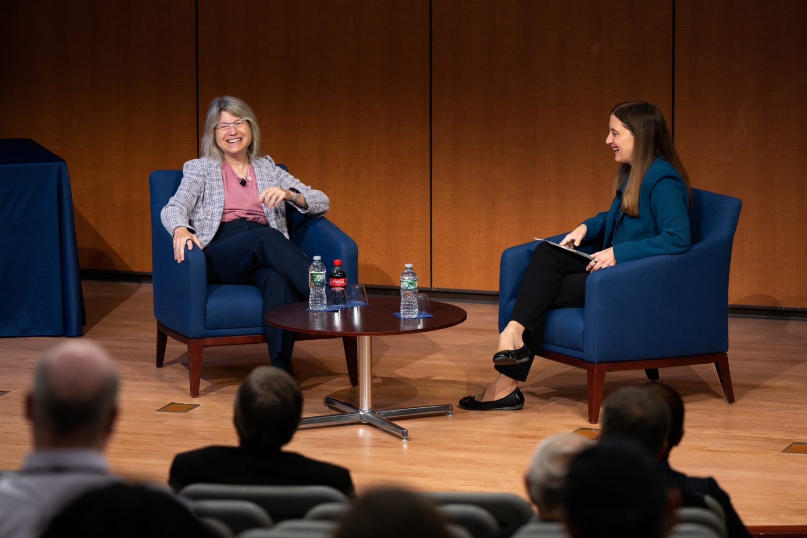 Sally Kornbluth and Melissa Choi sit in blue chairs on a wooden stage in Lincoln Laboratory's auditorium. 