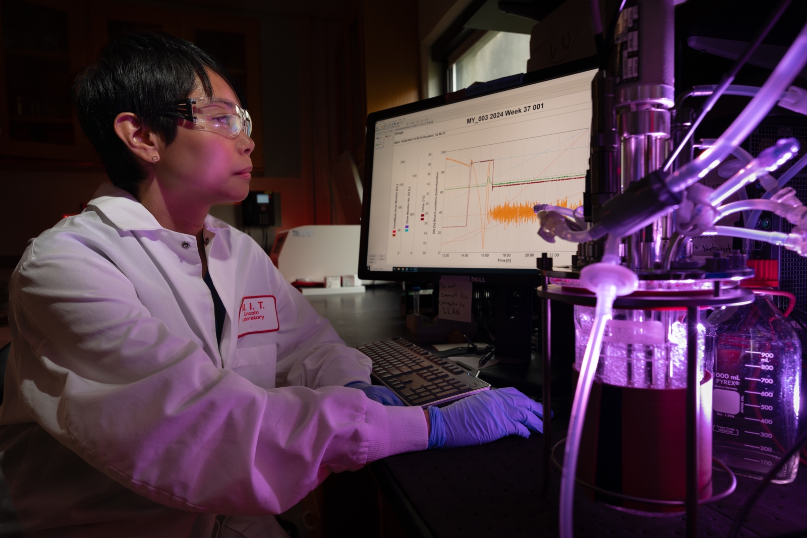 A researcher sits in front of a computer screen in a biotechnology lab. The screen shows data on a plot. 