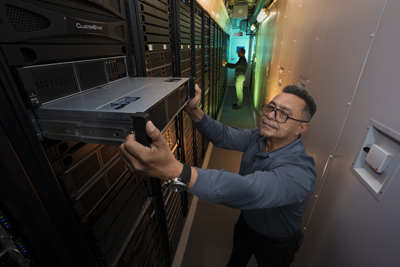 Standing within a narrow hallway containing racks of computer equipment, a system engineer pulls out a server (a black box about 2 feet long by 4 inches tall) off a rack to inspect it. 