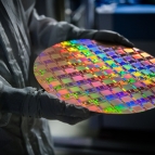 A person with a protective gown and gloves holds a large wafer of integrated circuits. the wafer is vividly multicolored.