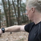 A man in black t-shirt looks at a smartwatch on his wrist, which is displaying a green circle with a thumbs up sign. The man is outside, with trees and leaves in the background.