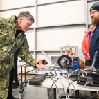 A military official looks at sensors on a table, across from a Lincoln Laboratory researcher. They are in large room, similar to a warehouse setting.  