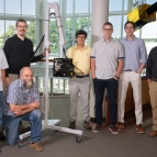 Seven individuals stand with a portable armature holding a laser-based ultrasound system.