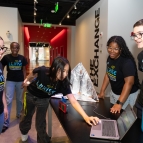 A group of LLRISE students wearing LLRISE shirts smile while preparing equipment inside of the MIT Museum.