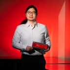 A photo of Jeffery Lim against a red background, holding a hardware platform (rectangle, about 4 by 12 inches). 